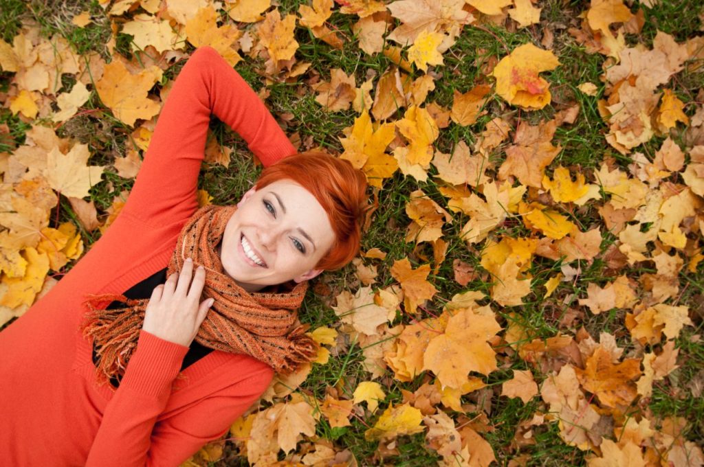 Smiling redheaded woman laying in colored fall leaves wearing a scarf